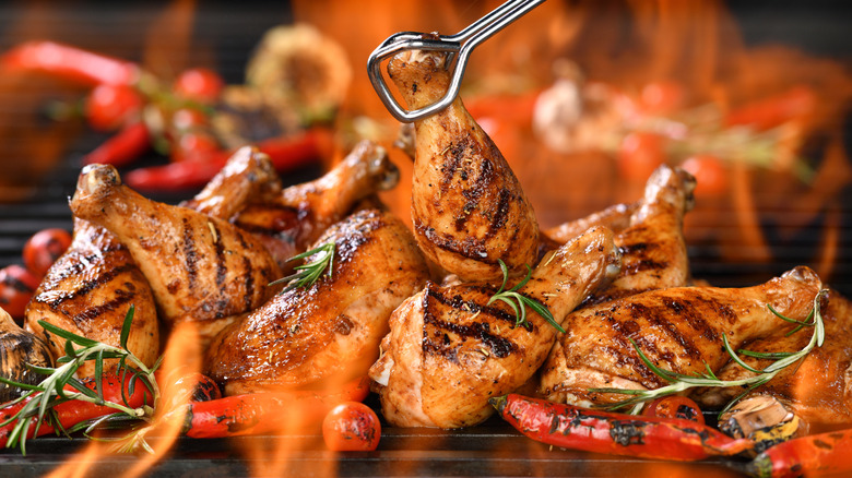 Chicken legs and thighs on a flaming grill with long peppers and rosemary. One chicken leg is being held up by a pair of tongs