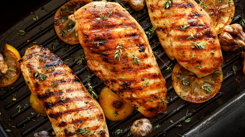Three chicken breasts with grill marks sitting on a barbecue grill with lemon, garlic, and thyme