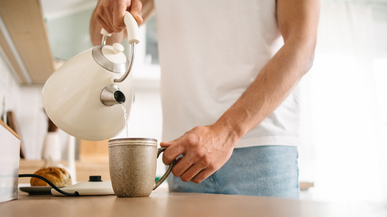 A man in a white T-shirt pouring water from a white kettle into a mug