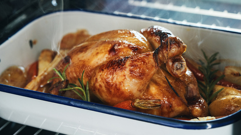 A whole roasted chicken in a roasting pan with fresh herbs and veggies
