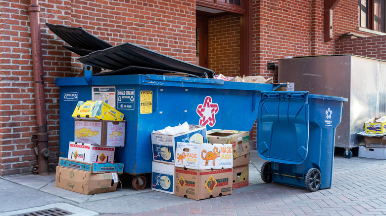 Blue dumpster and a blue trash can behind a brick building surrounded by boxes.