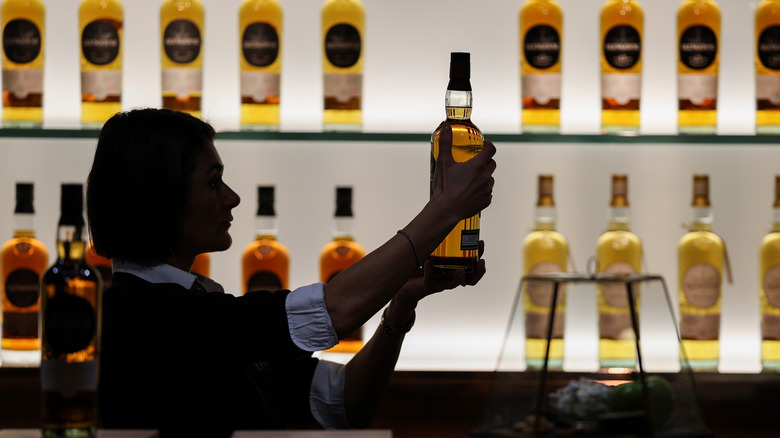 A woman closely admires a bottle of scotch whisky, against lit-up shelf holding bottles