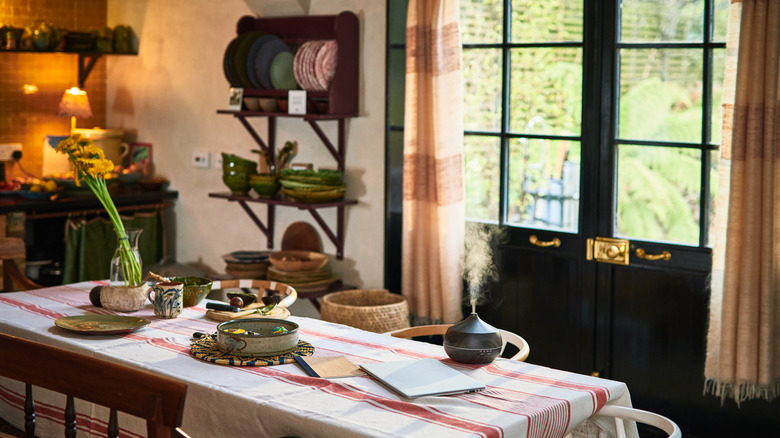 Country style kitchen with dark green door, beige curtains, and ceramic dishes and woven baskets on table and shelves