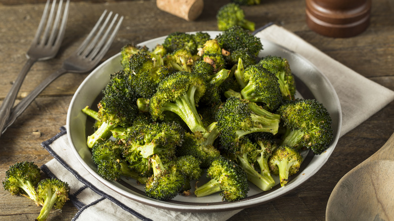 Roasted broccoli florets on a plate with a napkin, forks, a wooden spoon and more broccoli on the table.