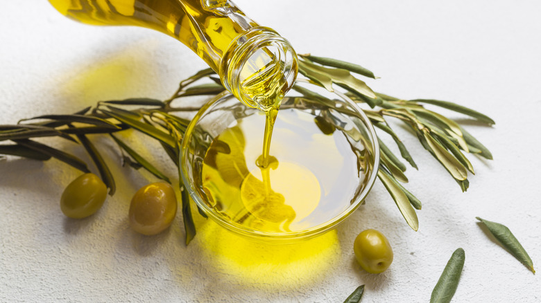 olive oil pouring from glass bottle into small glass bowl surrounded by olives and olive leaves