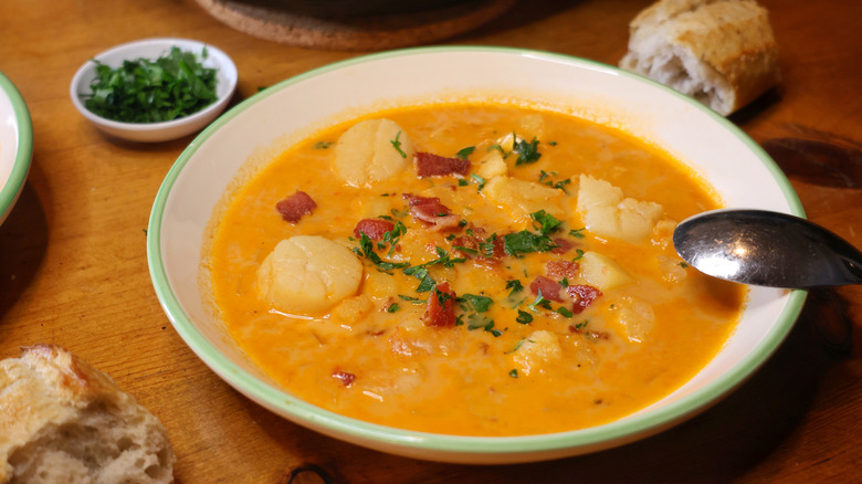 a bowl of Long Island seafood chowder with a spoon resting on the edge on a wooden table with a hunk of baguette and a small dish of minced parsley