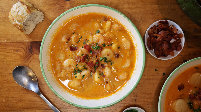 overhead view of a bowl of Long Island seafood chowder on a wooden table with a spoon, a piece of baguette, and a small dish of bacon bits