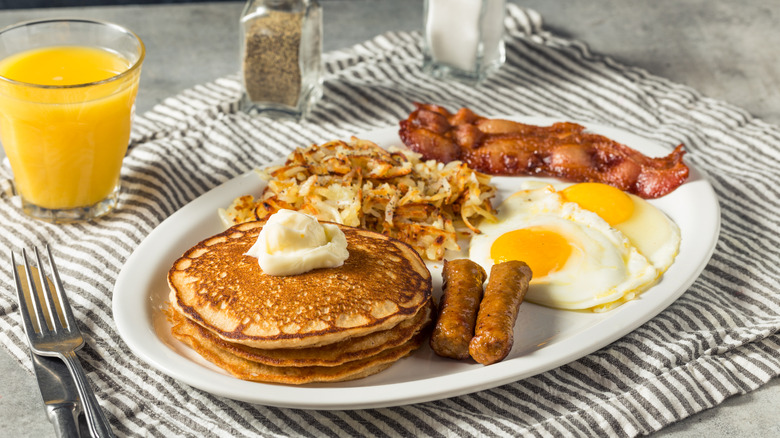 Pancakes, eggs, hashbrowns, and bacon on a plate beside a glass of OJ and salt/pepper shakers