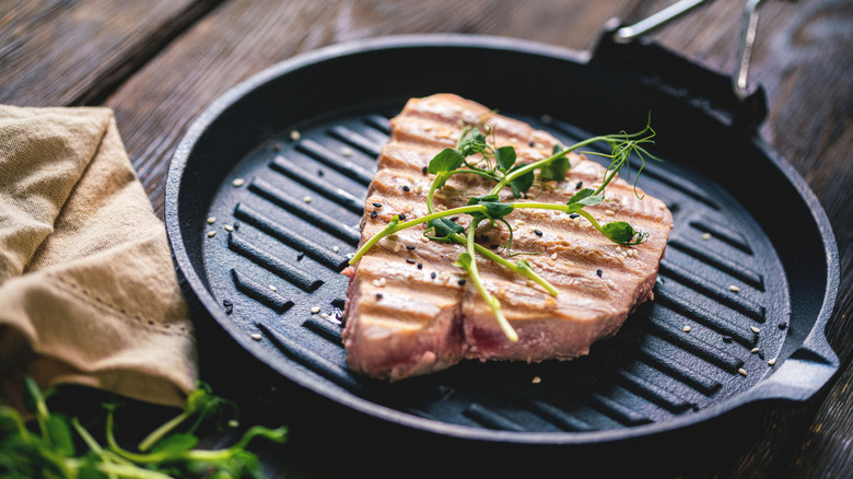 a grilled tuna steak topped with sesame seeds and microgreens in a cast iron grill pan on a wooden table