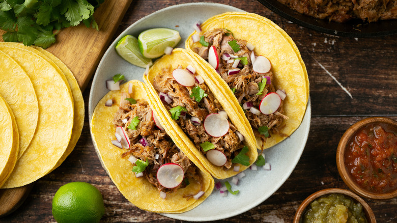 A plate of three carnitas tacos on corn tortillas with cilantro, red onion, radish, and lime wedges