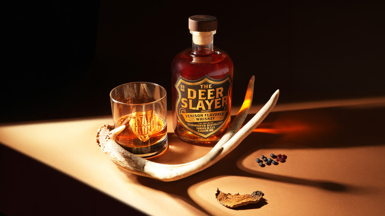 A bottle of The Deer Slayer Venison Flavored Whiskey beside a glass of whiskey an antler, and other natural items.