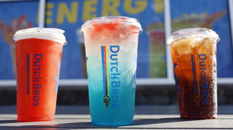 Dutch Bros red hot coffee cup, cup of blue and red energy drink, and iced coffee cup sitting on pavement outdoors