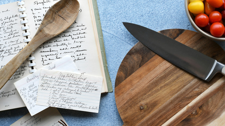 recipe book, wooden spoon, recipe cards, knife and board and bowl of tomatoes