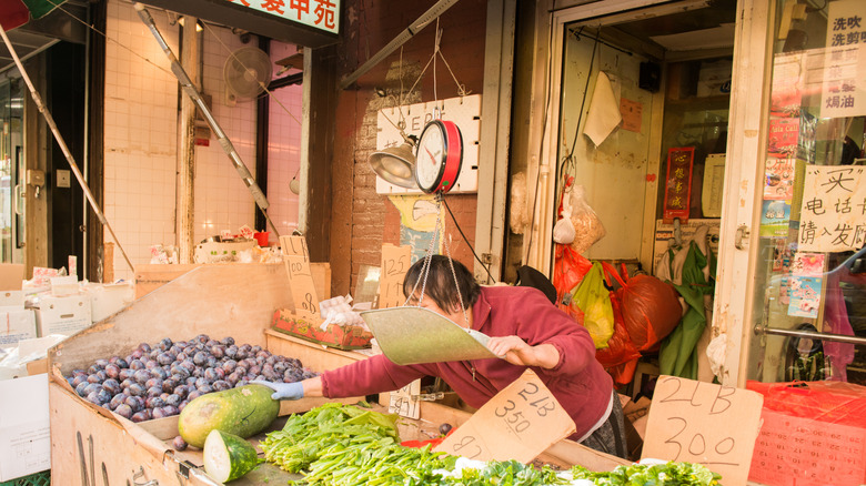 An outdoor stall in New York's Chinatown on Mott Street selling fresh produce, with an employee reaching for a large vegetable while holding a free-hanging scale