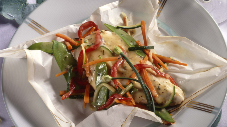 A baked chicken breast baked en papillote sitting on a plate covered in peppers, carrots, mangetout and a creamy sauce