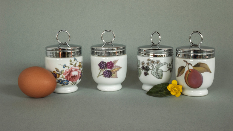 Four decorative porcelain egg coddlers on a kitchen counter next to an egg