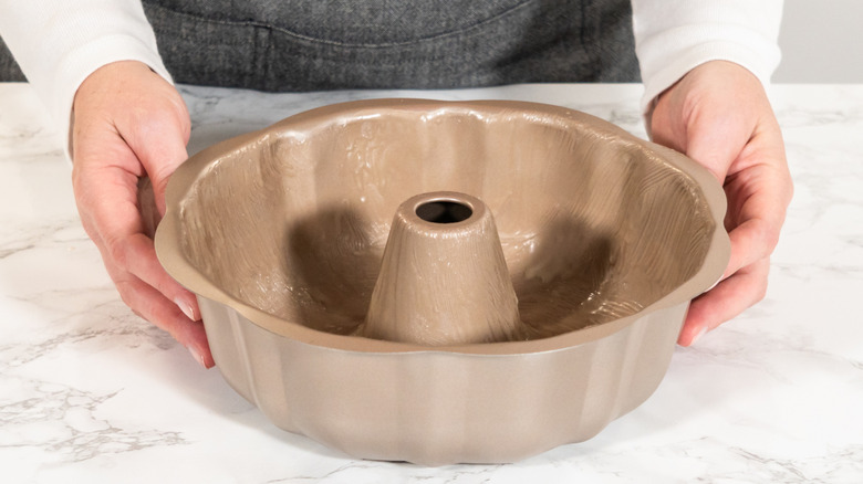 A woman in an apron holding a greased Bundt pan on a kitchen counter