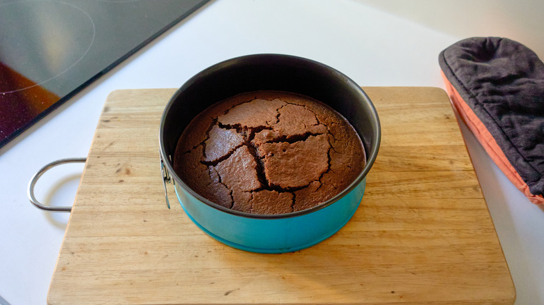 Domed chocolate cake with cracks on top in blue pan on wooden cutting board