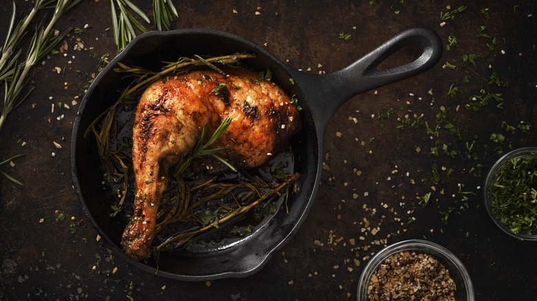 Roast chicken leg in a small cast iron skillet with herbs on a table scattered with various seasonings