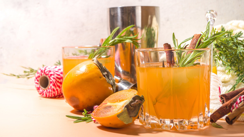 Two orange cocktails in tumblers, with a cocktail shaker, twine, cut persimmon, rosemary and cinnamon