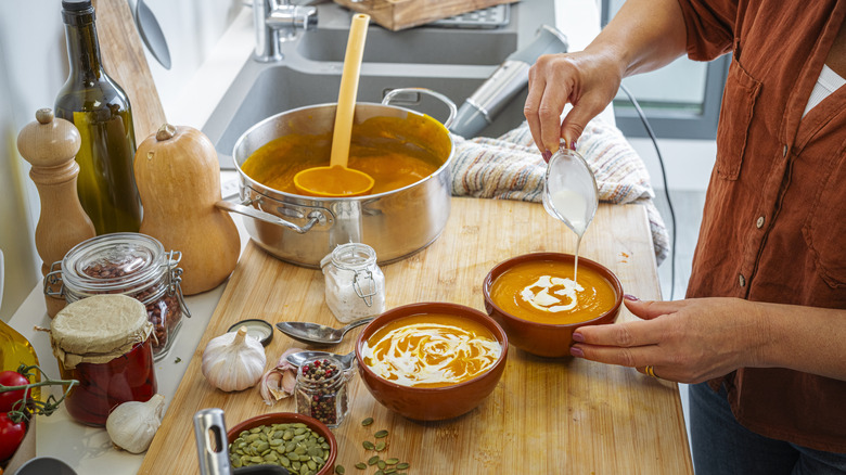 Person drizzling cream onto freshly-made bowls of soup on a cutting board in the kitchen