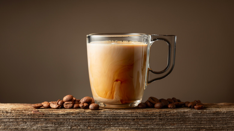 A flat white in a glass mug surrounded by coffee beans