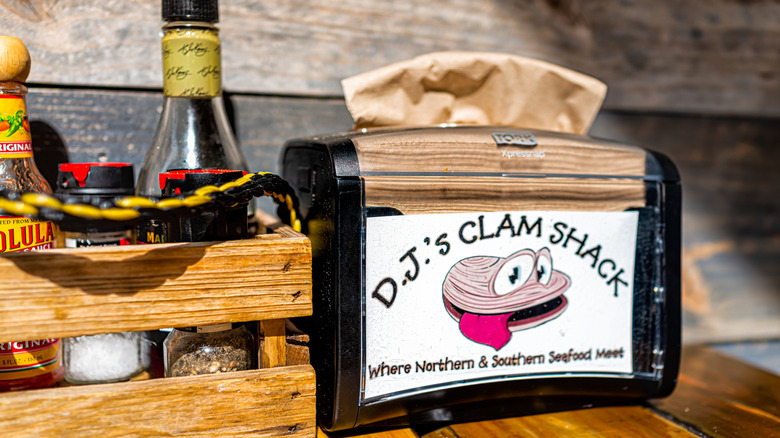 Napkin dispenser with the name and logo of D.J.'s Clam Shack, beside a box of sauces and seasonings