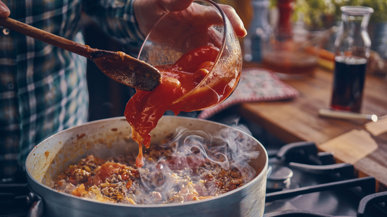 A person adding a bowlful of tomatoes to a pot of simmering spaghetti sauce