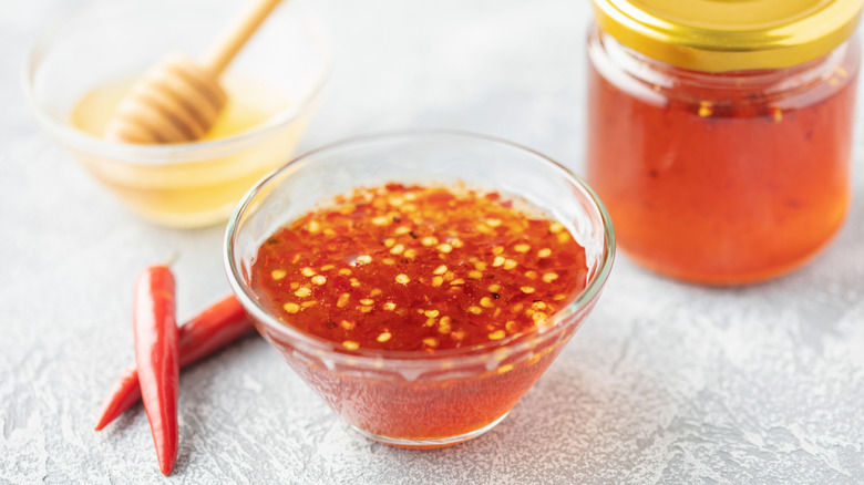 A bowl of hot honey with visible chili pepper seeds floating in it, a pair of red peppers on the table next to it, and honey in the background