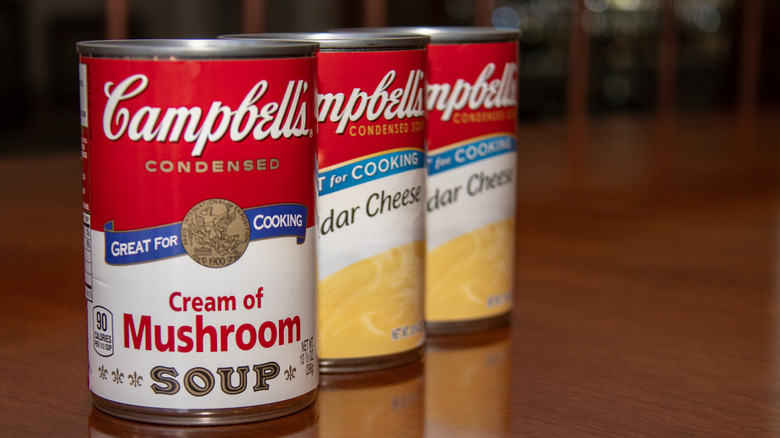 A can of Campbell's Cream of Mushroom soup on a table in front of two cans of Cheddar Cheese soup