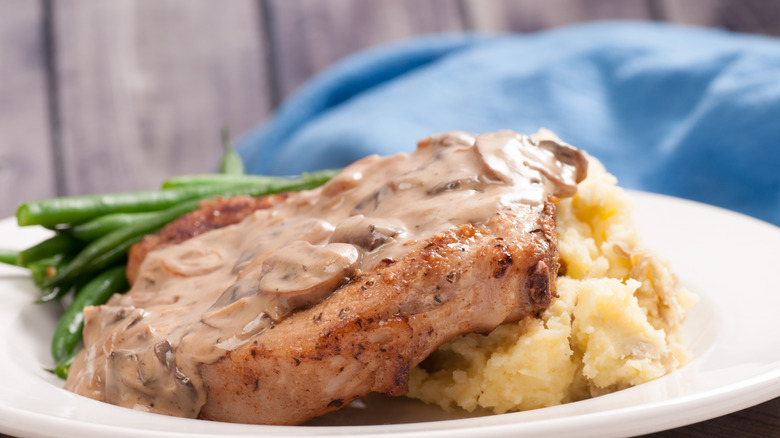 Pork chop resting on mashed potatoes ,with green beans in the background, on a white plate. The chop is covered in a mushroom sauce