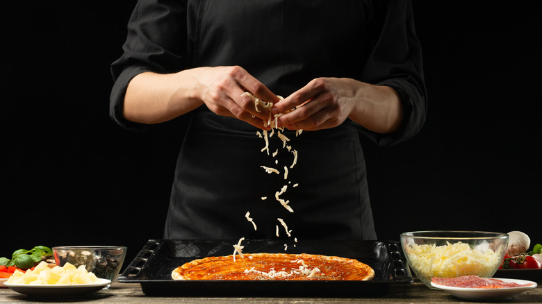 Chef sprinkling cheese onto a pizza base with sauce, toppings in bowls beside him