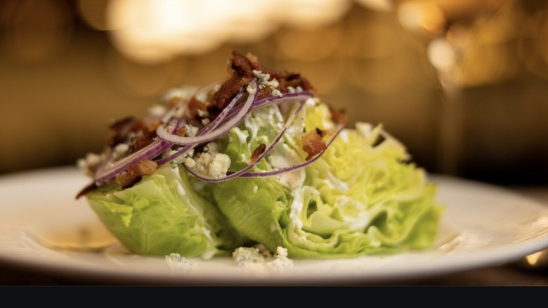 Delmonico's wedge salad with iceburg lettuce, red onions, bacon, and crumbled blue cheese on a white plate.