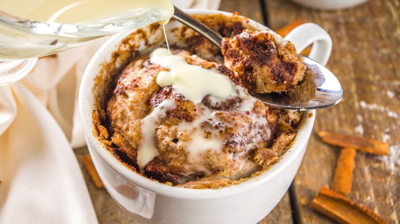 a cinnamon roll in a mug, a single bite displayed on a spoon, with sauce being poured over