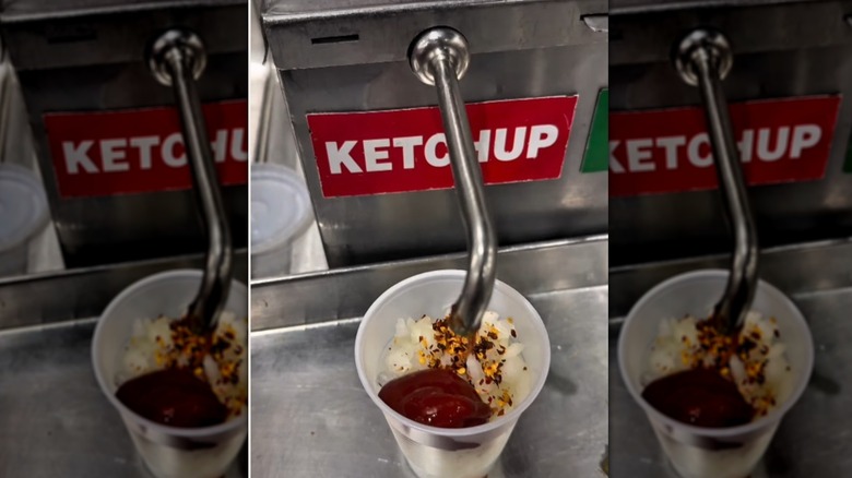 Plastic cup loaded with onions and red pepper flakes being filled with ketchup from the aluminum ketchup dispenser at a Costco