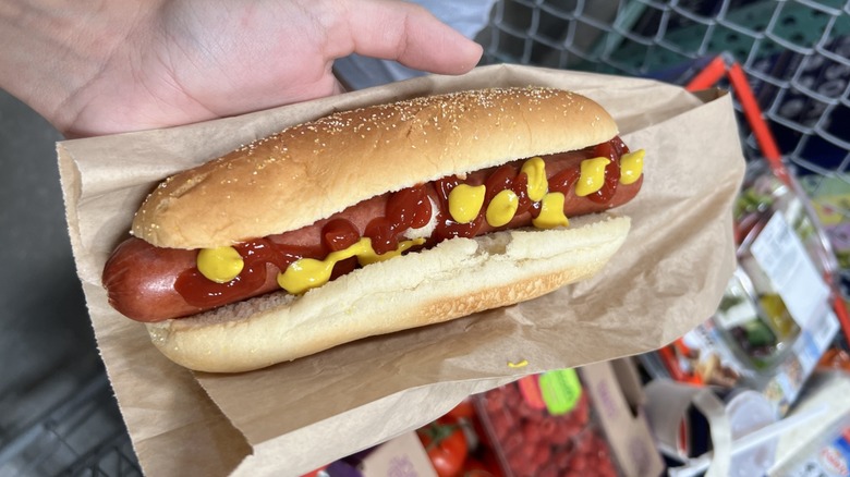 A person holding a Costco hot dog topped with mustard and ketchup, sitting on top of paper bag with a Costco shopping cart in the background