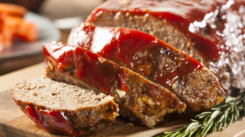 Sliced meatloaf with a ketchup glaze on a cutting board next to a sprig of rosemary