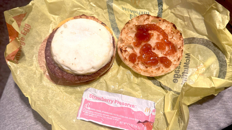 An Egg McMuffin with the top muffin removed showing egg and sausage on one side and strawberry preserves on the English muffin on the other side, all sitting atop a yellow wrapper