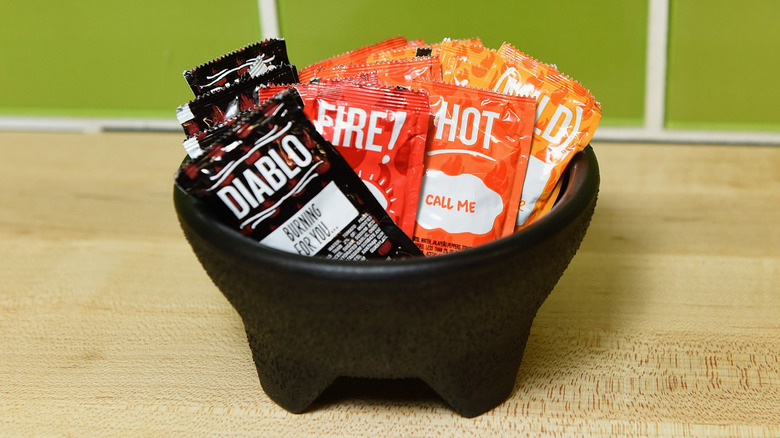 A bowl of Taco Bell sauces including Diablo, Fire, Hot, and Mild on a wooden table with a green tile wall behind