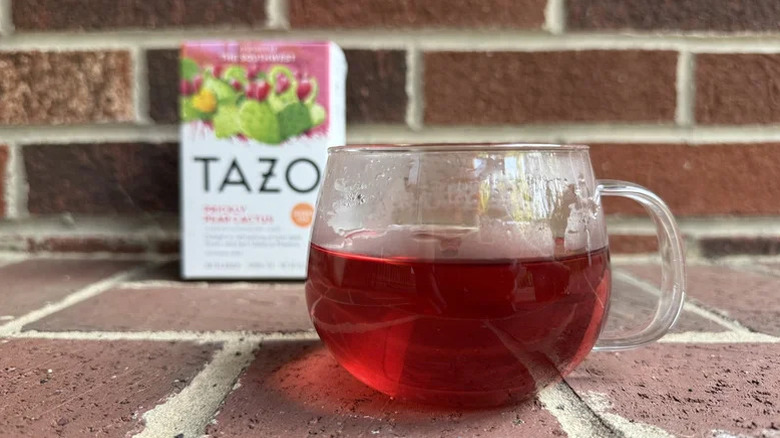 A clear mug of Tazo Prickly Pear Cactus Herbal Tea sits on a brick walkway with a box of the tea in the background.