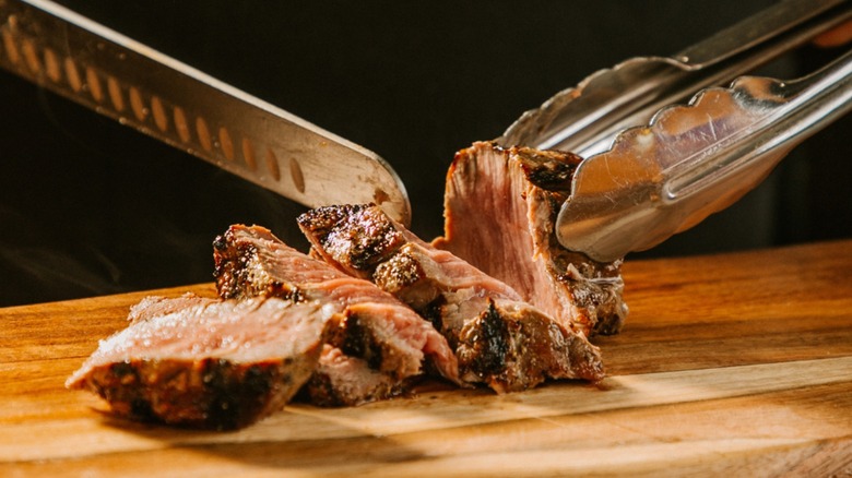 A knife cutting into a steak on a wooden cutting board while held in place by metal tongs