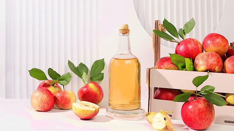 Apple cider vinegar in a glass container, surrounded by cut fresh apples and apples in a crate