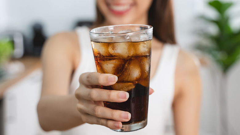 A smiling woman in a white tank top holding up a glass of soda with ice