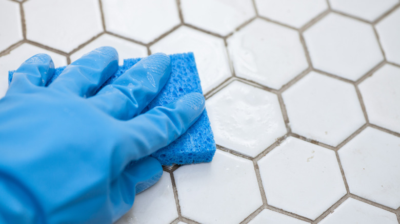 Gloved hand with a sponge cleaning dirty tile grout.