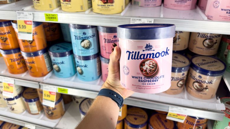 Hand holding a pink tub of Tillamook ice cream in front of a store fridge stocked with the brand's other flavors
