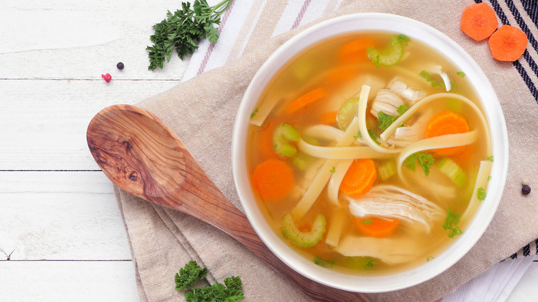 Bowl of chicken soup with noodles and carrots next to wooden spoon