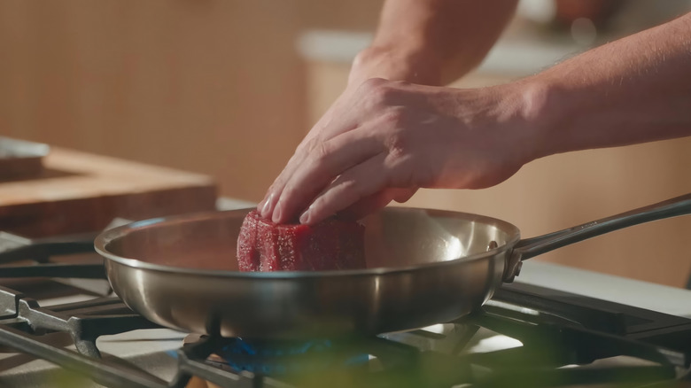A hand pressing down on a steak in a steel pan