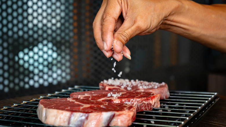 A hand seasoning raw steaks on a wire rack with salt