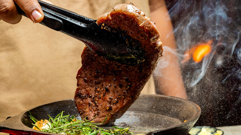 A person using tongs to flip a steak in a pan while cooking