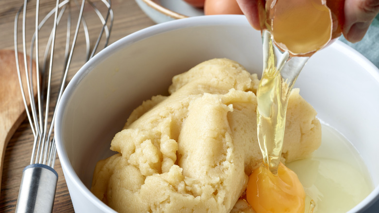 Person adding eggs to bowl of choux paste with whisk nearby